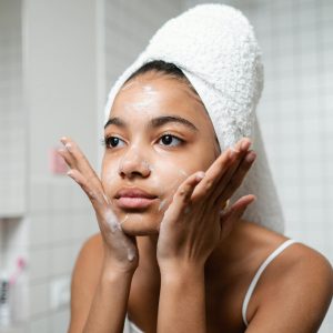 Woman with a towel wrapped around her head applies skincare in bathroom.